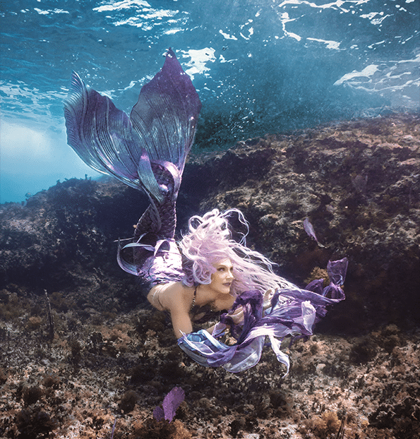 Real-life mermaid with a violet tail swimming above a rocky seabed